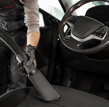 Une personne passe l'aspirateur à l'intérieur d'une voiture à l'aide d'un tuyau, en se concentrant sur les sièges. Elle porte des gants et une tenue de travail. Le volant est visible, ce qui suggère la propreté