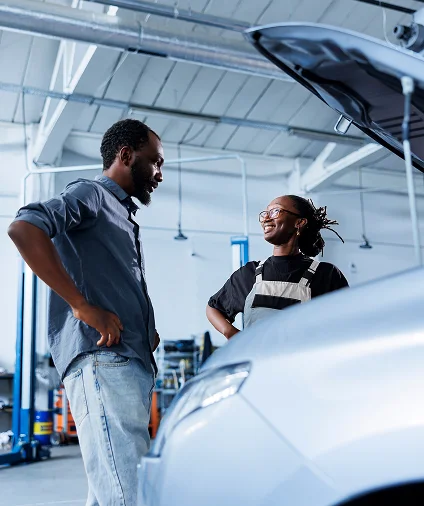 Un mécanicien et un client sourient tout en discutant d'une voiture dans un atelier de réparation automobile lumineux. L'ambiance est conviviale et professionnelle